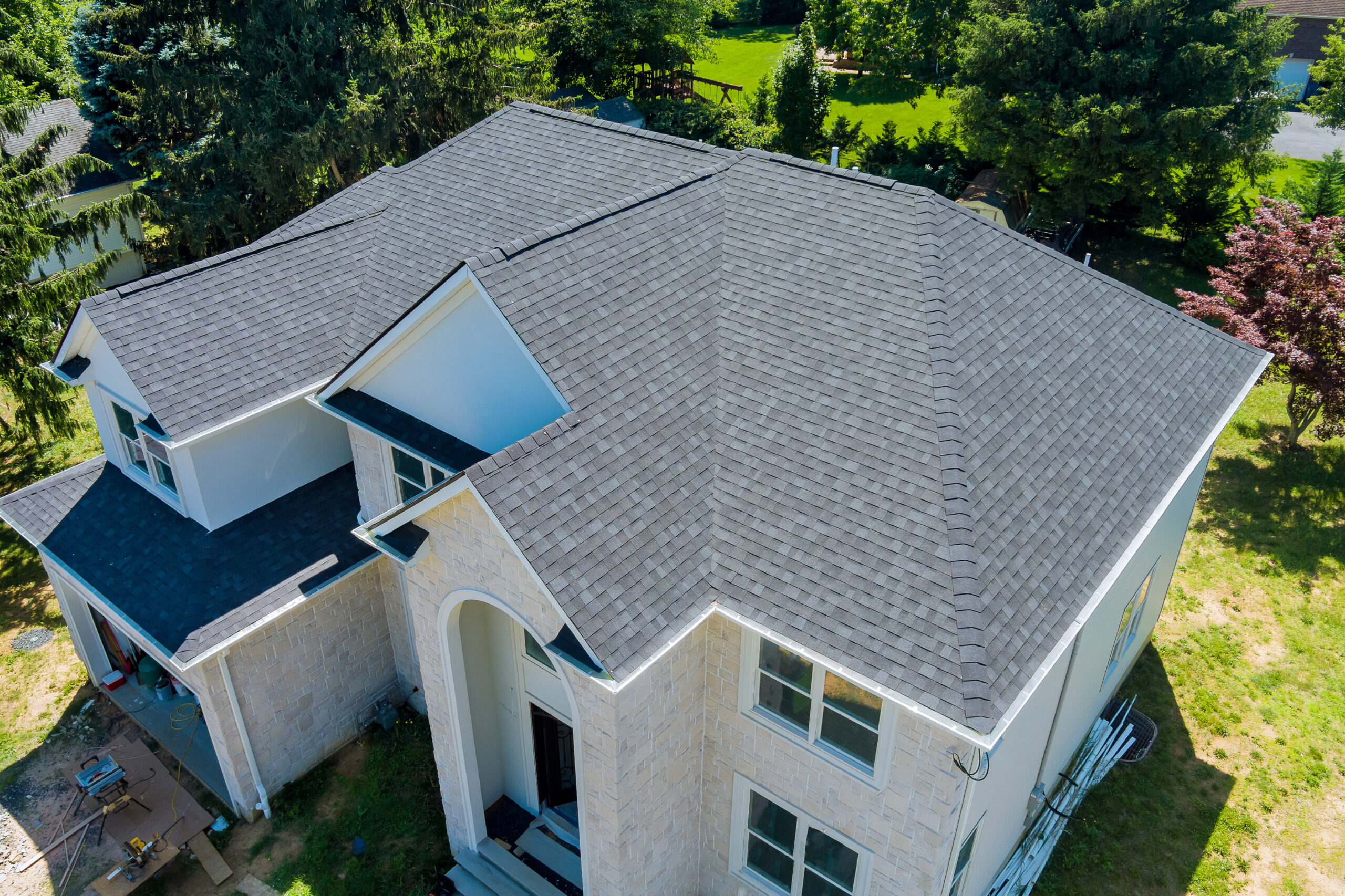 Aerial view of a new gray shingle roof on a two story stone home, with trees and a green lawn around it